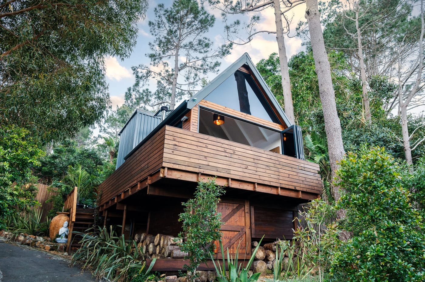 The A-frame cabin on stilts at dusk, its glazed gable glowing above the log store