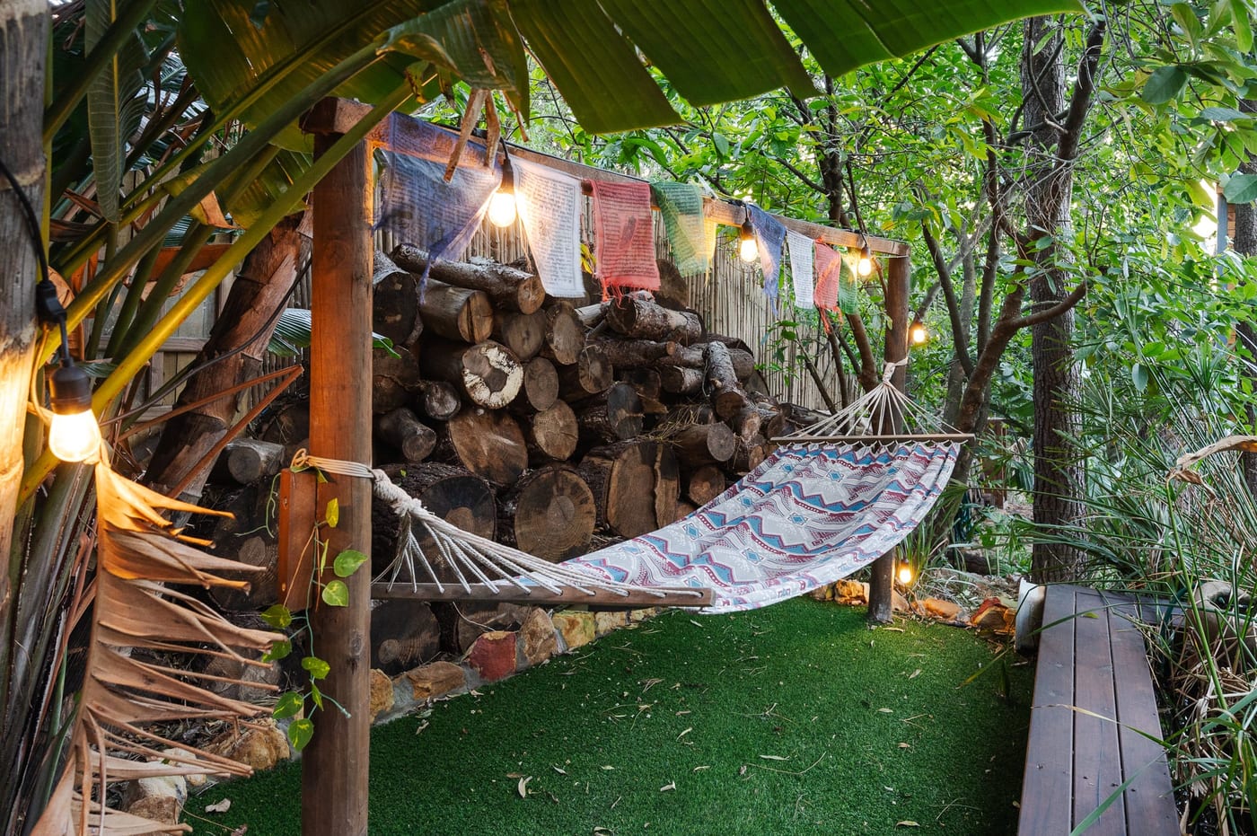 A hammock strung before a stacked log wall, prayer flags and festoon lights above
