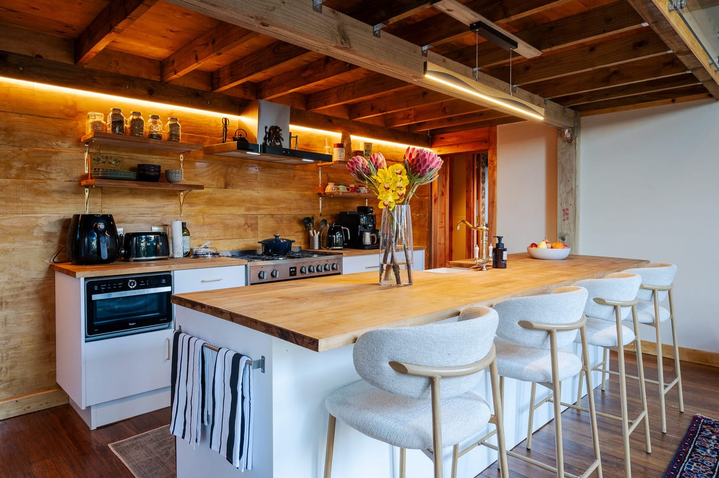 Four upholstered stools at the timber-topped island, shelves lit above