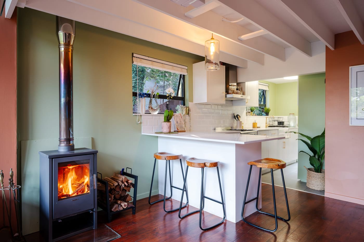 Live-edge stools at the peninsula, the stove lit and firewood stacked alongside