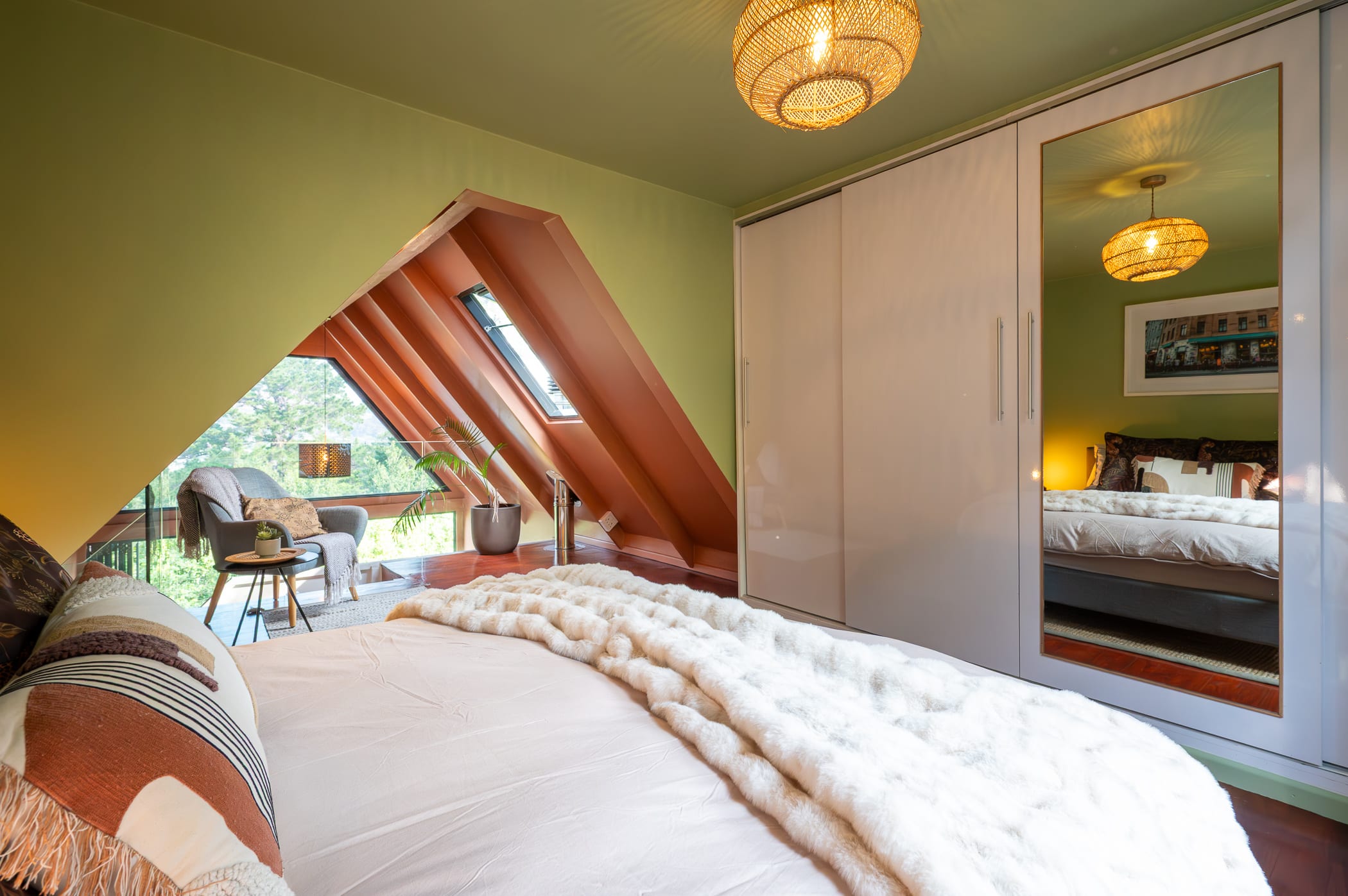 The loft bedroom under the A-frame gable, bed made up beside a seating nook looking into the trees