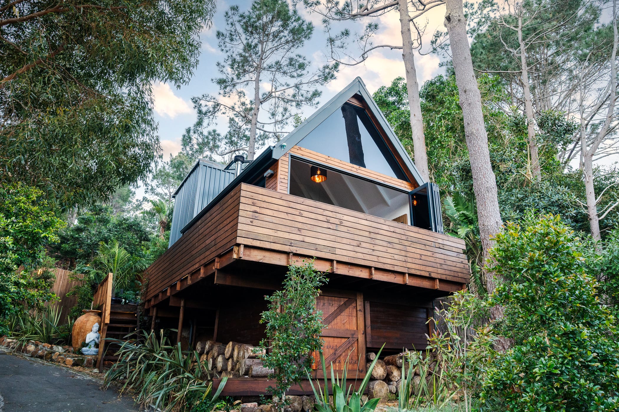 The A-frame cabin on stilts among the pines at dusk, its glazed gable glowing above the log store